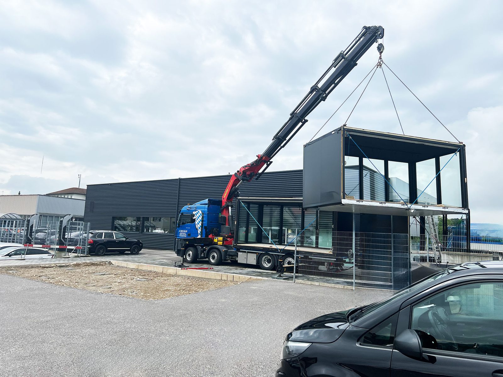 Ladekran eines blauen Lkw hebt ein schwarzes Modul mit Glasfront an Ketten vor einer dunklen Gewerbehalle